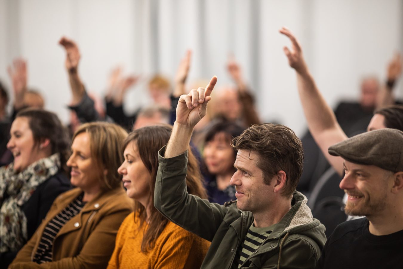 crowd of happy-seeming seated people, some with their hands up.