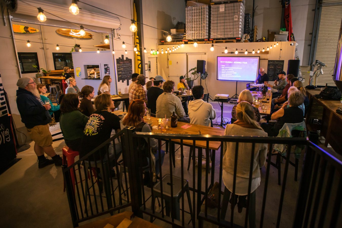 Crowd of seated and standing people watching a presentation, shot from the back of the room.