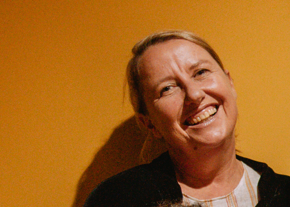 Ali Buckley head and shoulder shot with Ali smiling and hair tied back, wearing a striped peach and white t-shirt and black cardigan in front of a yellow background.