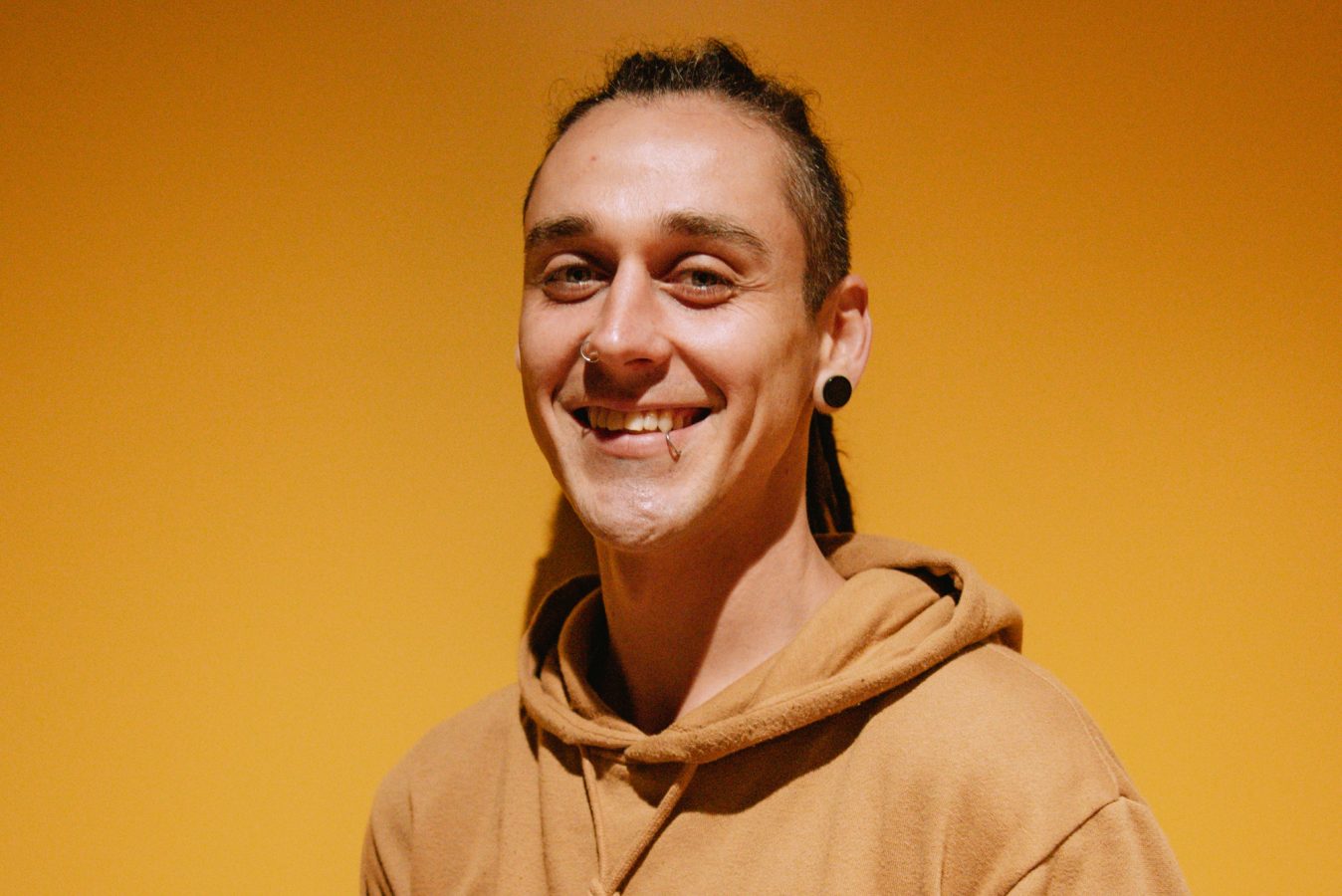 Head and shoulder shot of Kayne Mills with long hair tied back, wearing a yellow hoodie that somewhat matches the yellow background.