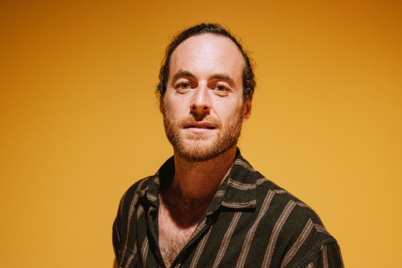 Head and shoulder shot of Sam Rees with dark hair tied back, wearing a striped striped shirt in front of a yellow background.