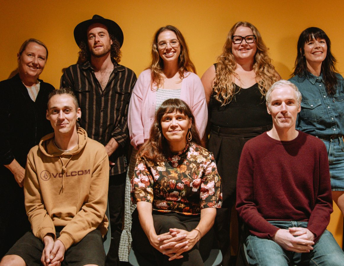 Eight MusicNSW staff members pose in front of a yellow back drop