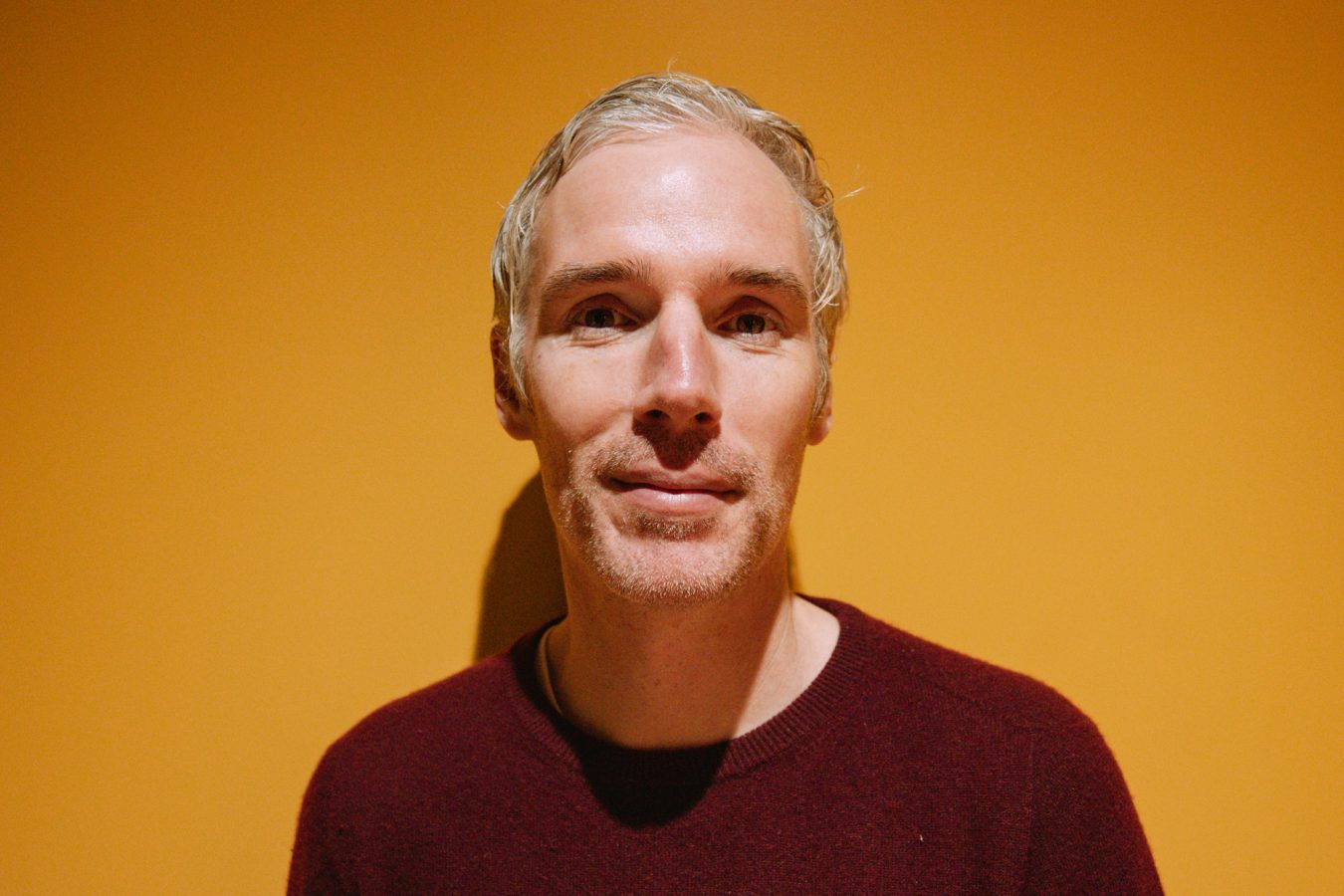 Head and shoulder shot of Dave with short grey hair wearing a dark red crew jumper standing in front of a yellow background.