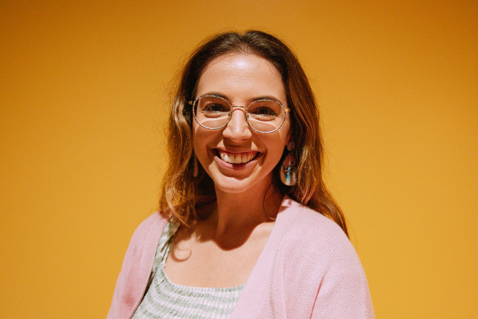 A smiling sophie Jones stands in front of a yellow background. Sophie has dark blonde hair wearing glasses and a pink cardigan.