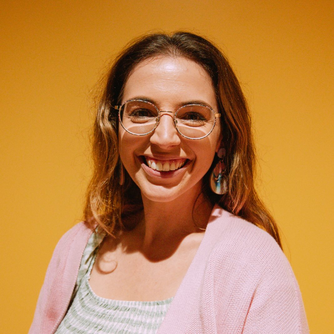 A smiling sophie Jones stands in front of a yellow background. Sophie has dark blonde hair wearing glasses and a pink cardigan.